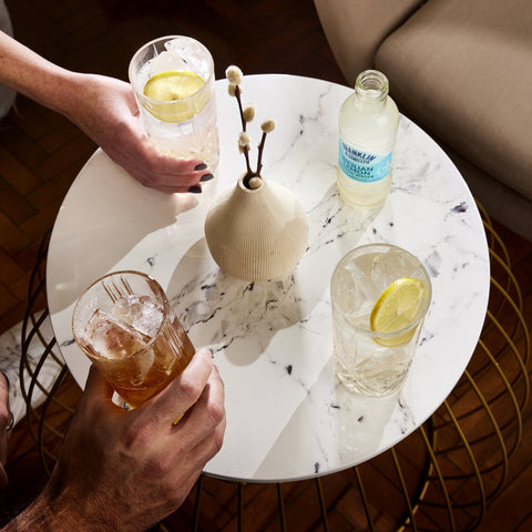 A social setting with a person holding a glass of Franklin & Sons Sicilian Lemon Tonic Water with ice and a lemon slice, another glass on the table, and a bottle of tonic in the background.