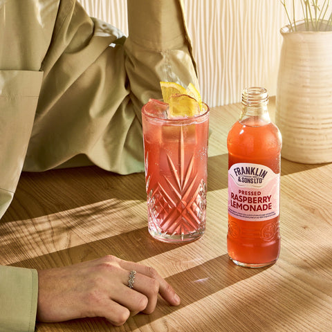 A bottle of Franklin & Sons raspberry lemonade next to someone with a glass of the lemonade, Franklin & Sons are premium soft drinks. 