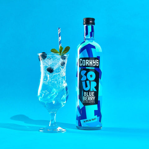 A bottle of Corky's sour blueberry schnapps next to a blue drink on a bright blue background. 
