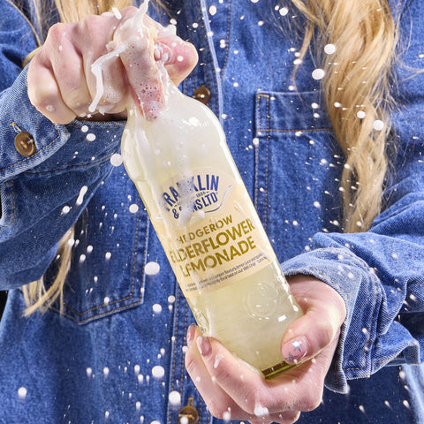 Someone holding a bottle of Franklin & Sons hedgerow elderflower lemonade as the lemonade splashes out the glass, Franklin & Sons have premium soft drinks. 