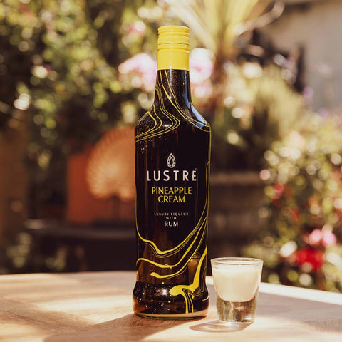 A bottle of Lustre Pineapple Cream Rum, a vegan cream liqueur, with an alcoholic shot of the cream liqueur next to the bottle. 