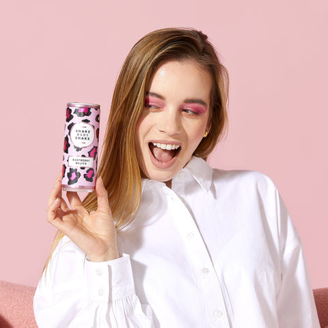 A person holding a can of Shake Baby Shake Raspberry Mojito cocktail with a pink background.