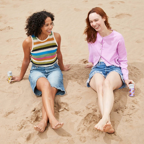 Two women sitting on a beach drinking All Shook Up Canned Cocktails in flavours Pina Colada or Passion Fruit Martini. 