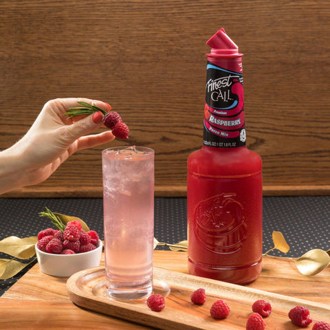 Bottle of Finest Call raspberry next to a pink drink and surrounded by raspberries on a wooded board. 