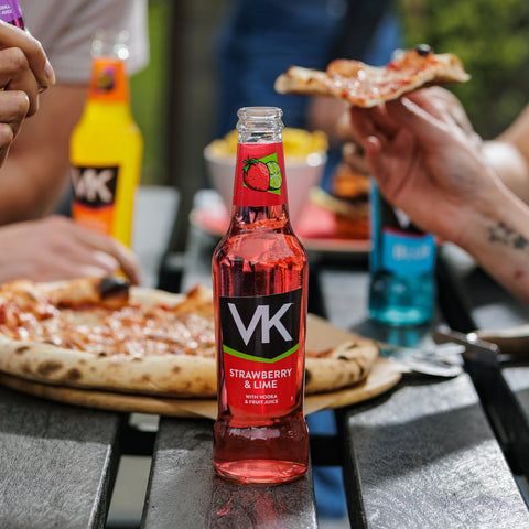 A bottle of VK strawberry and lime in the front focus with other VK ready to drink alcopops in the background with people eating pizza. 
