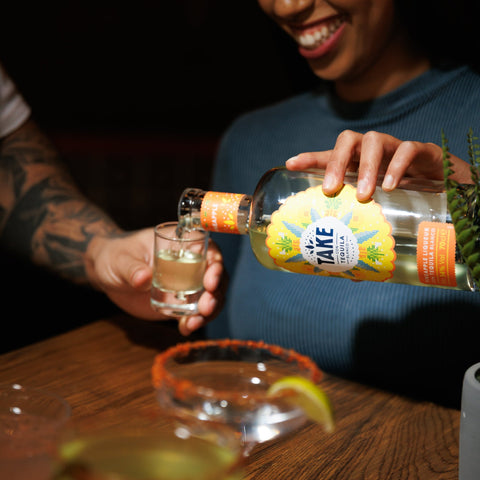 Person pouring a shot from a bottle labelled Take Tequila into a glass on a wooden table.