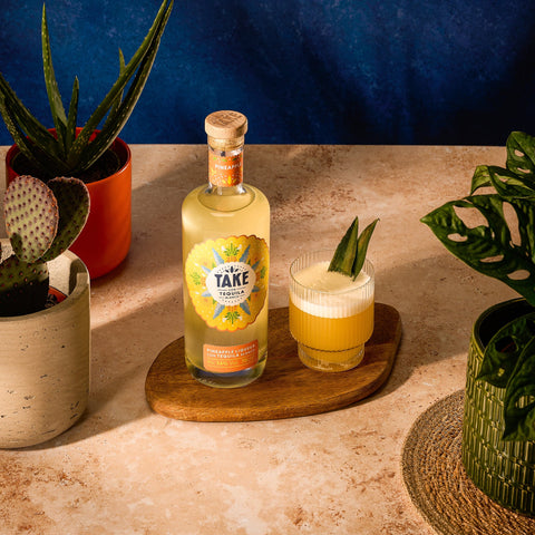 Bottle of Take tequila with a glass of pineapple-themed cocktail on a wooden coaster, surrounded by plants.