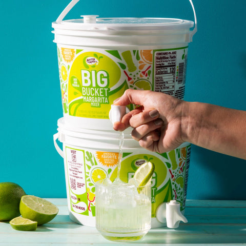 A person's hand squeezing a lime into a glass, with Master of Mixes 'BIG BUCKET MARGARITA' in the background, alongside limes and a tequila bottle.