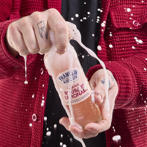 Someone holding a bottle of Franklin & Sons rhubarb lemonade as it splashes out of the bottle, Franklin & Sons offer premium soft drinks. 
