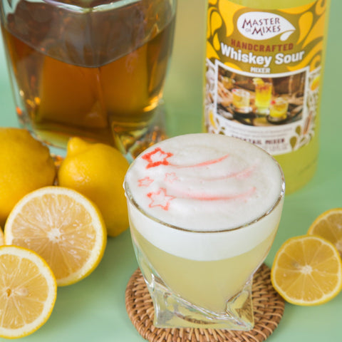 Glass of whiskey sour with lemons and a bottle of Master of Mixes Whiskey Sour mix on a green background