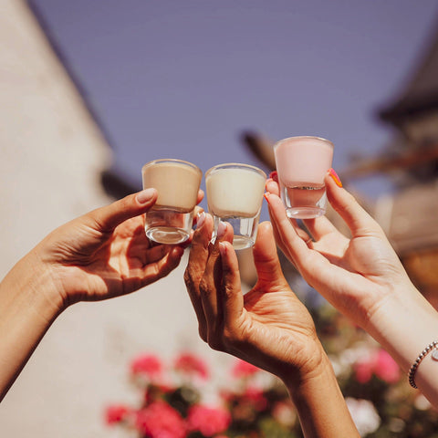 Three people holding alcoholic shots of Lustre vegan cream liqueur in each flavour, Caffe Latte Vodka Cream, Pineapple Rum Cream and Strawberry Tequila Cream. 