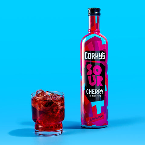 A bottle of Corky's sour cherry schnapps next to a glass of schnapps and ice on a bright blue background. Corky's is great for easy shots and cocktails. 