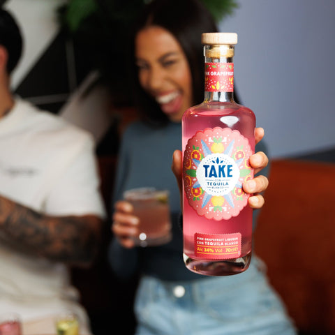 Person holding a bottle of pink grapefruit Take tequila with a blurred background of people and a table.