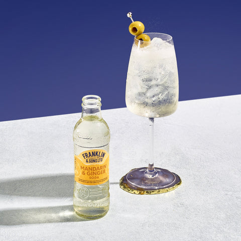 A bottle of Franklin & Sons Mandarin & Ginger Soda next to a tall glass filled with ice and a slice of orange, garnished with a green olive on the side.