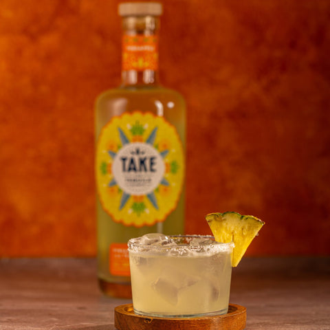Bottle of 'Take' with a cocktail and pineapple wedge on a wooden surface against an orange background