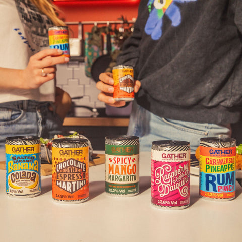 Five cans of GATHER cocktails lined up on a table with two people in the back holding another two cans of GATHER. 