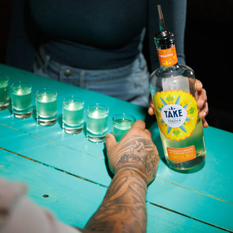 Person holding a bottle of Take Tequila with glasses on a teal table.