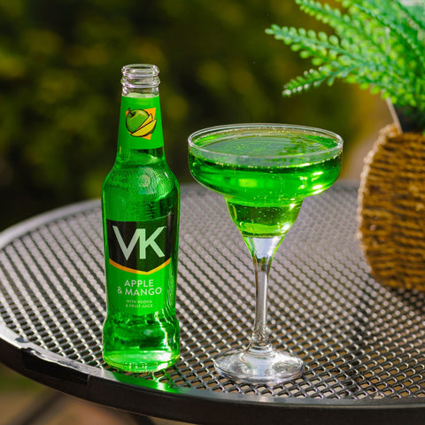 A bottle of apple & mango VK next to a cocktail glass of apple and mango VK on a table outside. 