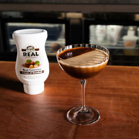 A hazelnut cocktail that has been made using Re'al Cream of Hazelnut Syrup, with the bottle of Re'al syrup placed next to the cocktail. 