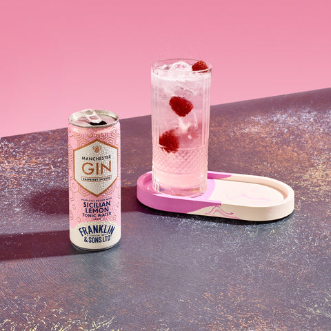 A ready to drink can of Manchester raspberry gin & Franklin & Sons Sicilian lemon tonic water next to a glass that is filled with the drink and raspberries. 