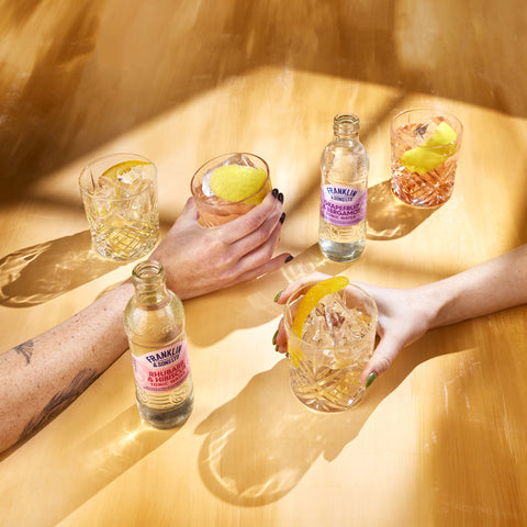 A image of Franklin & Sons Rhubarb Tonic Water with Hibiscus and Franklin & Sons grapefruit & bergamot tonic, showing bottles and glasses with the tonic on a yellow background. One can see hands holding the glasses and lemon slices as garnish.