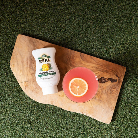 Squeezey bottle of real pineapple syrup on a wooden cutting board with a pink drink and an orange slice on a green surface
