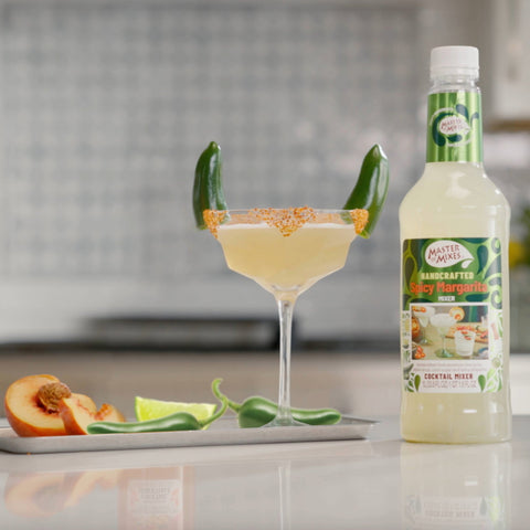 A bottle of Master of Mixes spicy margarita with a decorated cocktail and chilli's, limes and peach in the background in a kitchen. 