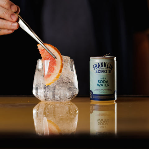 A glass of Franklin & Sons 1886 Soda Water with ice and a slice of citrus being garnished, next to a can of the premium product.