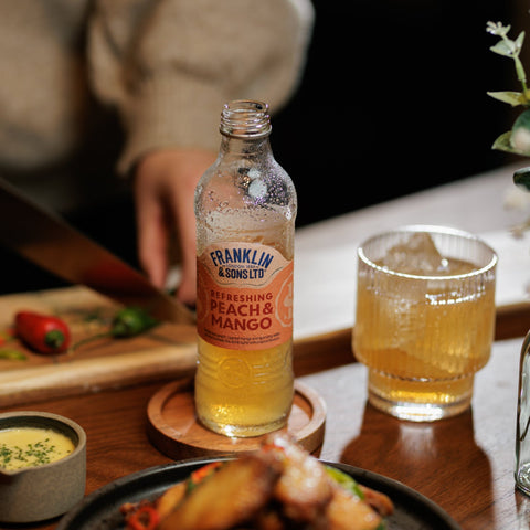 Bottle of Franklin & Sons Peach Mango premium soft drink and a  glass on a table with food.