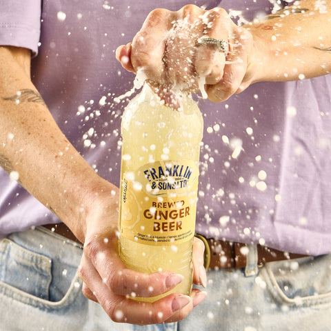 A person holding a bottle of Franklin & Sons Brewed Ginger Beer, a perfect mixer, as the contents are being poured out, creating a splash.