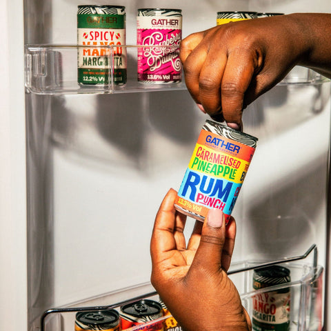 A person holding and opening a can of GATHER caramelised pineapple rum punch with GATHER cocktail cans in the background in the fridge door.