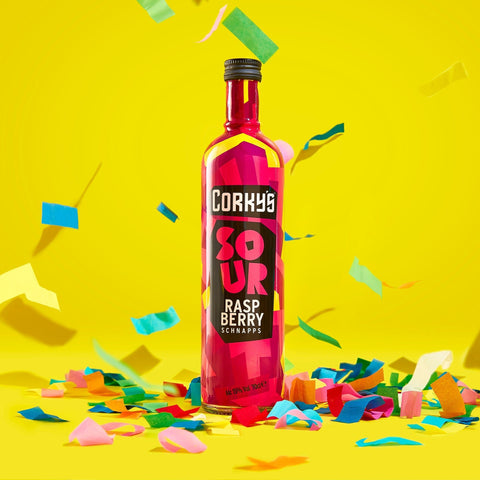 A bottle of Corky's sour raspberry schnapps on a bright yellow background with multicoloured  confetti. 