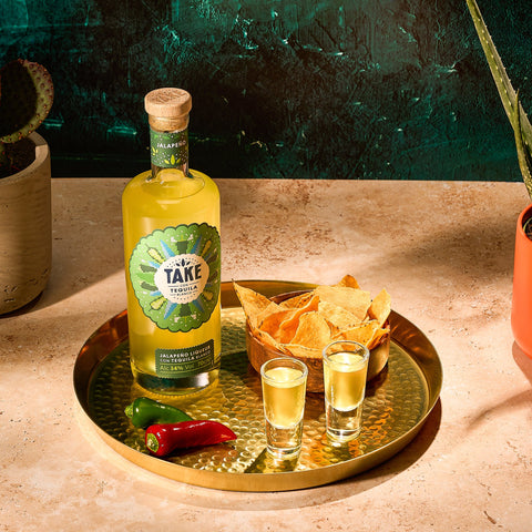 A bottle of Take Tequila jalapeno liqueur next to a bowl of crisps, chilis and two shots of a gold tray. 