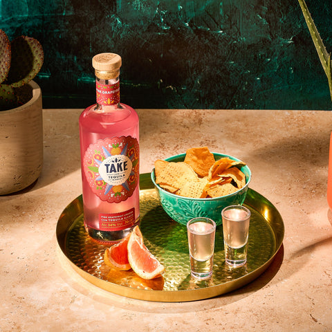 A bottle of Take Pink Grapefruit Tequila Liqueur with a colorful label, alongside a bowl of chips and two shot glasses on a golden tray, garnished with a slice of pink grapefruit and a piece of lime.