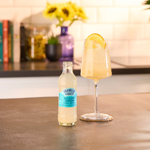Bottle of Franklin & Sons Sicilian Lemon soda next to a glass with lemon slice on a wooden surface.