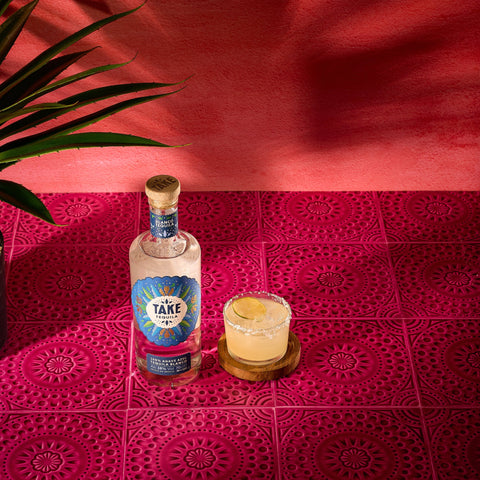A bottle of Take Tequila Blanco, made with 100% agave azul, with a margarita cocktail next to the bottle.