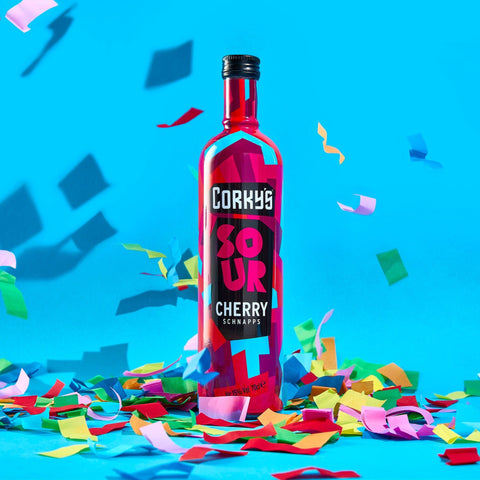 A bottle of Corky's sour cherry schnapps on a bright blue background with multi-coloured confetti. 