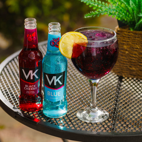 Two bottles of VK vodka mix alcopop in flavours black cherry & original blue next to a glass that is filled with VK. 