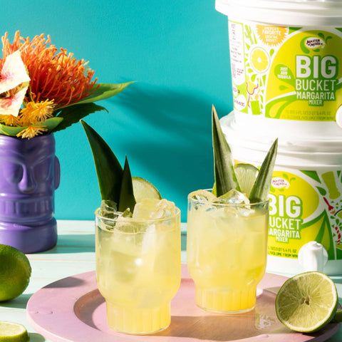 Two margarita drinks with garnish and limes with Master of Mixes margarita big buckets in the background. 

