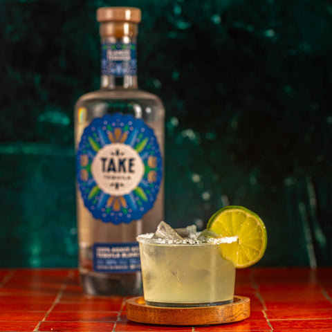 Bottle of 'Take' tequila with a cocktail garnished with a lime wedge on a red surface.