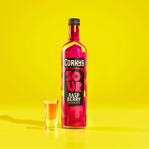 A bottle of Corky's sour raspberry schnapps on a yellow background next to a shot glass with pink liquid. 