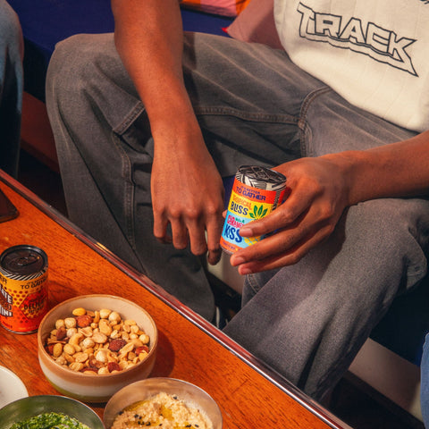 A person holding a can of GATHER with another can of GATHER and snacks on the table in front of them. 