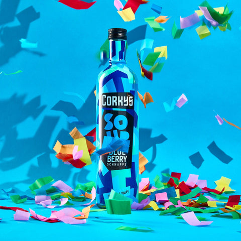 A bottle of Corky's sour blueberry schnapps on a bright blue background with multi-coloured confetti. 