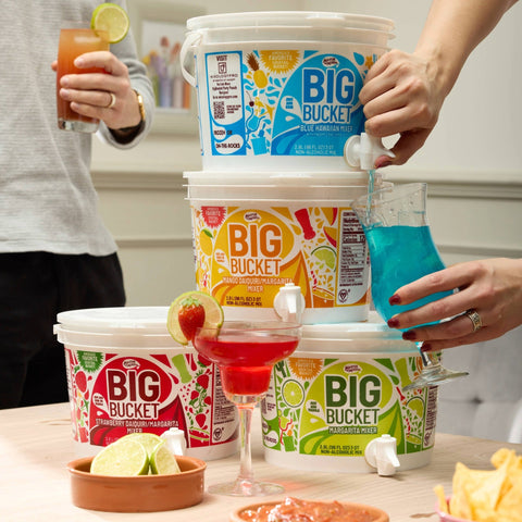 Person pouring a drink from a Master of Mixes Big Bucket Blue Hawaiian Mixer. With other Big Bucket Cocktail Mixers stacked on top of one another, in a party setting. 