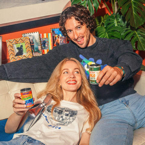 Two people sitting on a sofa with a plant in the background holding cans of GATHER cocktails. 