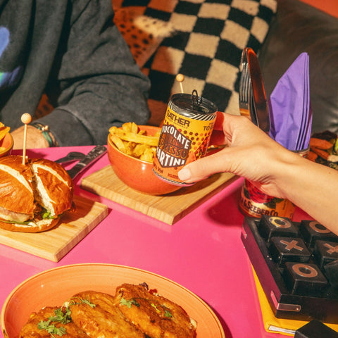 Someone holding a can of GATHER chocolate honeycomb espresso martini, with food on the table in the background. 