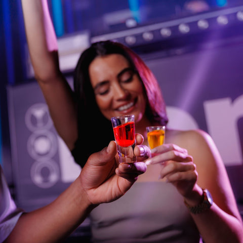 A girl holding a shot of Corky's sour schnapps with someone else holding another Corky's shot. 