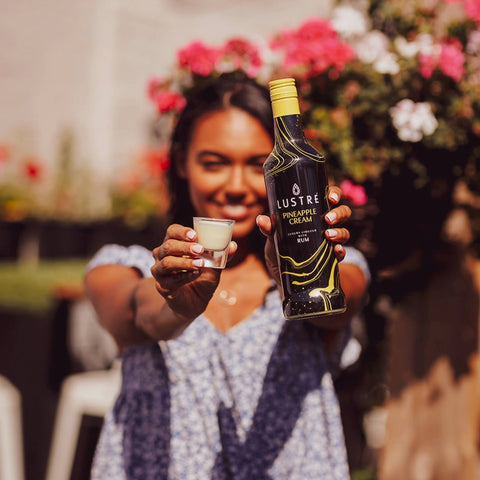 A woman holding a shot of Lustre Pineapple Cream Rum, with the Lustre bottle in the other hand. 