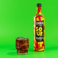 A bottle of Corky’s Sour Mango Schnapps with a colorful label, next to a glass filled with the schnapps over ice.