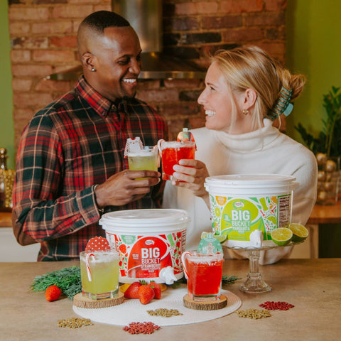 Two smiling people with drinks with two  aster of Mixes strawberry and margarita big buckets in front of them. 